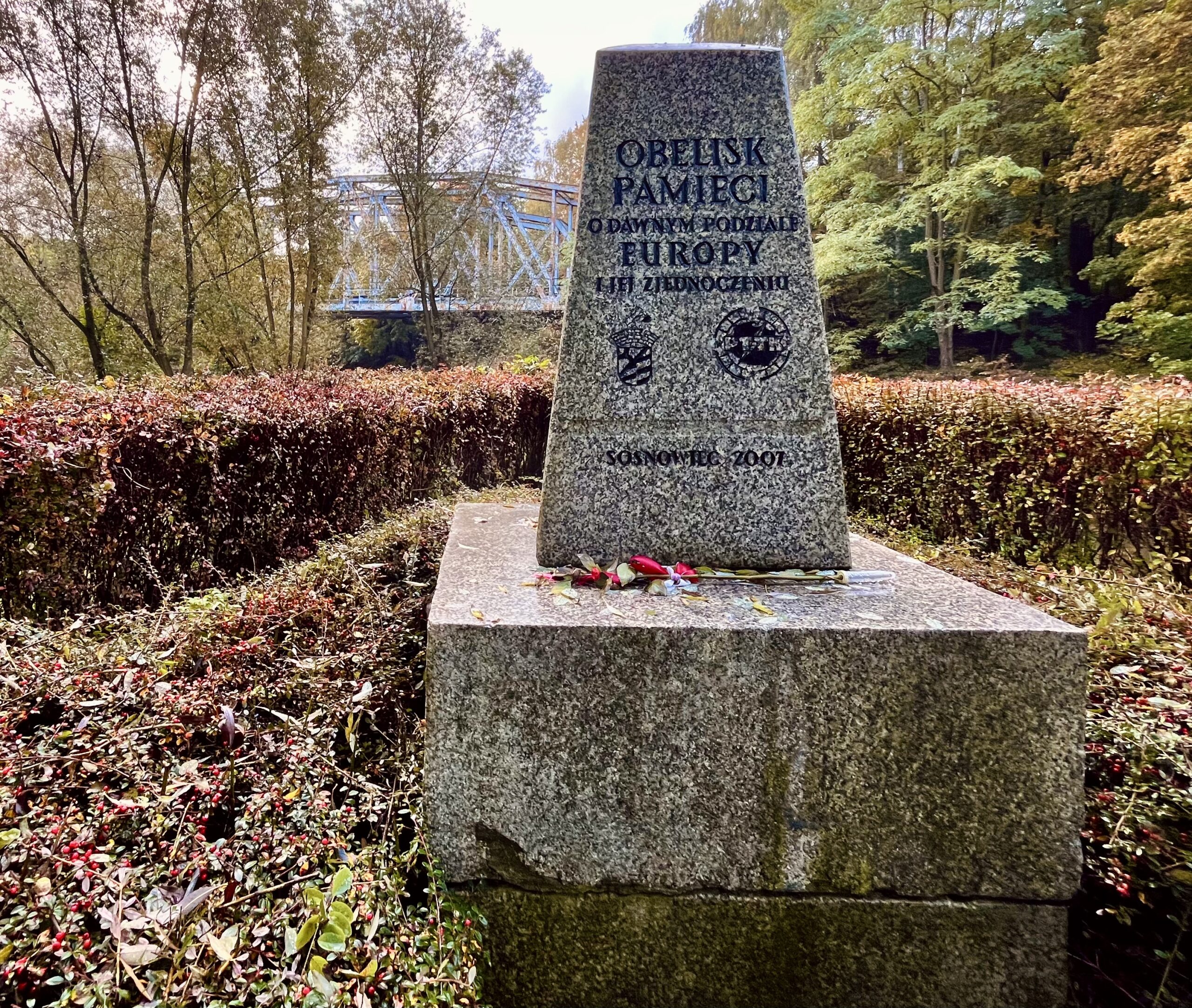 Granitowy obelisk upamiętniający dawny podział Europy i jej zjednoczenie, wzniesiony w 2007 roku w rejonie Trójkąta Trzech Cesarzy w Sosnowcu. W tle widoczny stalowy most kolejowy nad Przemszą w otoczeniu jesiennych drzew.