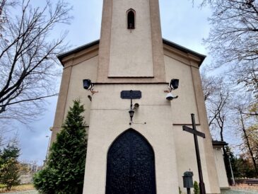 Fasada kościoła Najświętszego Serca Pana Jezusa w Sosnowcu, tzw. Kościółka Kolejowego. Na zdjęciu widoczna jest frontowa wieża z neogotyckim portalem, czarnymi ryflowanymi drzwiami oraz otoczeniem z drzewami, krzyżem i zniczami ustawionymi przy wejściu.