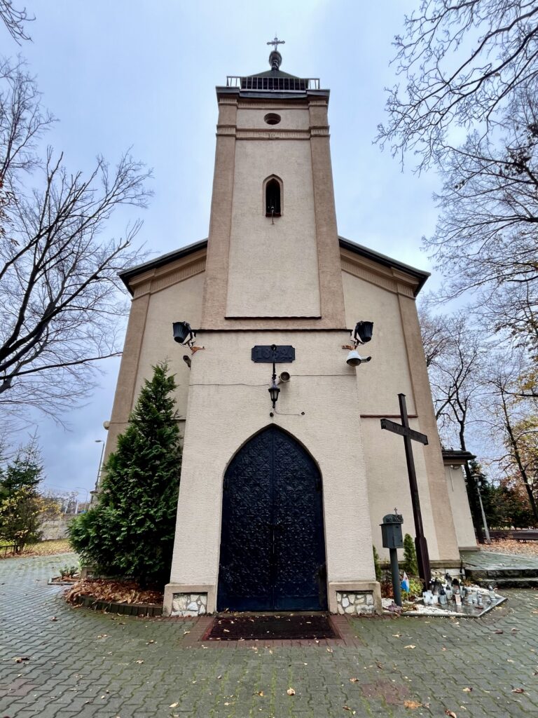 Fasada kościoła Najświętszego Serca Pana Jezusa w Sosnowcu, tzw. Kościółka Kolejowego. Na zdjęciu widoczna jest frontowa wieża z neogotyckim portalem, czarnymi ryflowanymi drzwiami oraz otoczeniem z drzewami, krzyżem i zniczami ustawionymi przy wejściu.
