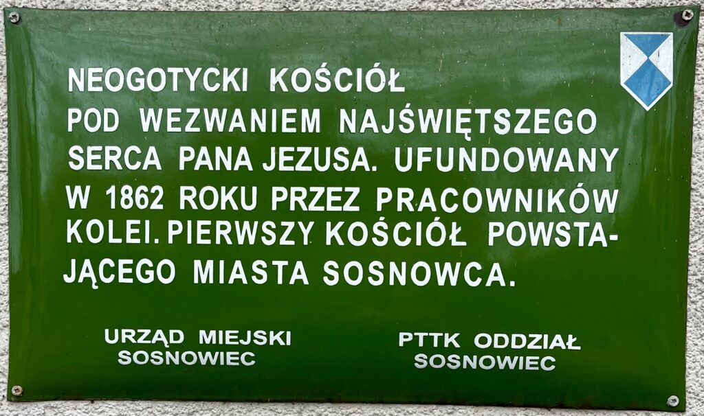 Zielona tablica informacyjna na elewacji kościoła Najświętszego Serca Pana Jezusa w Sosnowcu z białym napisem o historii świątyni oraz logotypami Urzędu Miejskiego i PTTK